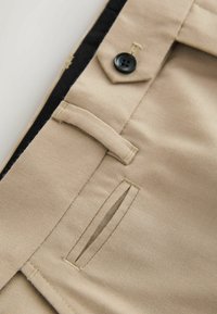 Beige Hosen mit glatter Textur, ausgestattet mit einem schwarzen Knopf, dezenten Nähdetails und seitlichen Taschenöffnungen.