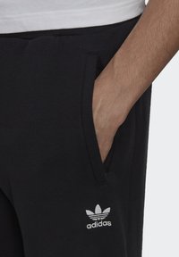 Čierne tepláky s bočnými vreckami, mäkkou textúrou a vyšívaným bielym logom Adidas na ľavom boku.