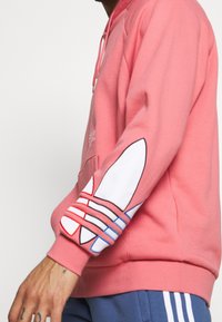 Ljusrosa hoodie med en känguruficka, ribbade muddar och en stor vit och färgad randig logga på vänster ärm.