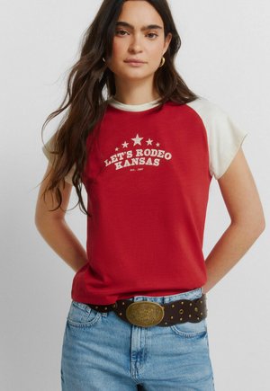 Vrouw met lang donker haar die een rood-wit T-shirt draagt met de tekst "LET'S RODEO KANSAS", een blauwe spijkerbroek en een bruine riem met een grote gouden gesp.