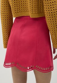 Primo piano di una gonna rossa con orlo scallop e aperture a goccia, indossata con un top di maglia giallo senape con un motivo a buchi.
