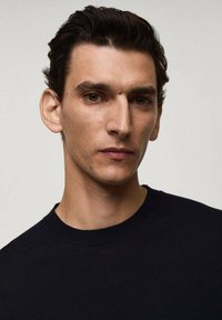Maglione nero a maglia con collo rotondo, indossato da una persona. Lo sfondo è neutro, concentrandosi sui tratti del viso e sulla texture dei capelli.