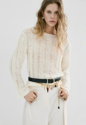 Mujer con cabello rubio ondulado que lleva un suéter de punto suelto color crema, pantalones blancos, cinturón negro y cadena dorada con dijes, anillos y pendientes.