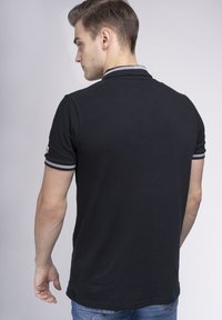 Camisa polo negra con cuello y mangas a rayas grises, diseño ajustado, textura suave y un detalle de logo en el lateral, combinada con jeans azules.