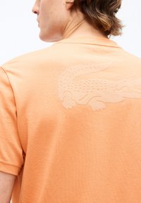 Άτομο με πορτοκαλί πουκάμισο που έχει υφασμένο λογότυπο κροκόδειλου στην πάνω πλάτη, γυρισμένο ελαφρώς στο πλάι, δείχνοντας το λαιμό και τα μαλλιά.