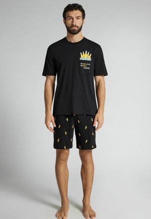 Mężczyzna stojący boso, ubrany w czarny T-shirt z butelkami piwa i napisem na kieszeni oraz dopasowane czarne szorty z żółtymi nadrukami butelek piwa.