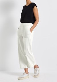Svart kortärmad topp i kombination med vita vida byxor med fickor. Modellen bär ljusa sneakers med beige detaljer. Ren, minimalistisk design.