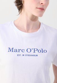 Balta kokvilnas T-krekls ar zilām drukātām uzrakstam "Marc O'Polo EST. STOCKHOLM." Klasisks apaļas kakla izmērs un mīksta tekstūra.