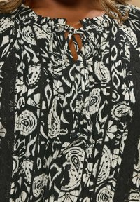 Svart och vit mönstrad blus med nyckelhål och knytkrage, texturerad tyg och plisserad design, med knappar framtill.