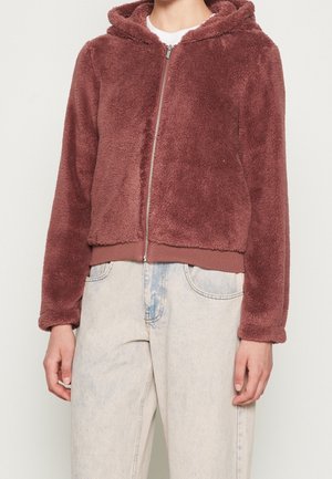 Personne portant un sweat à capuche marron en tissu duveteux avec fermeture éclair, et un jean taille haute délavé clair, debout devant un fond uni.