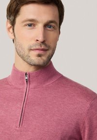 Maglione zip-up di un tenue rosa con colletto alto, realizzato in un morbido tessuto a maglia. Presenta una leggera texture e un dettaglio in metallo sulla zip al collo.