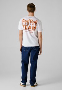 Bijela majica kratkih rukava s velikim narančastim natpisom "Red Bull Racing TEAM" na leđima, u kombinaciji s plavim trapericama i ležernim cipelama.