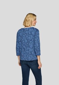 Blaue gemusterte Bluse mit Dreiviertelärmeln, ausgestattet mit einem strukturierten Design und einem runden Ausschnitt. Kombiniert mit dunkelblauen Jeans.