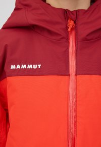 La veste Mammut rouge et rouge foncé présente une capuche, une fermeture éclair à l'avant et un logo blanc. Le tissu a une texture lisse avec un design coloré.