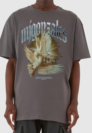 Person iført en grå t-shirt med to hvide duer og ordet "mignonsalles" i stiliseret skrifttype over dem.