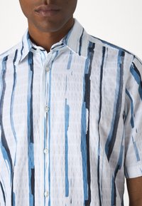 Koszula z krótkim rękawem w kolorze białym z pionowymi paskami w odcieniach niebieskiego i czarnego. Posiada kołnierzyk i zapinany z przodu na guziki. Materiał o fakturze.