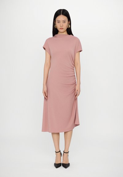 Knælang, blush pink kjole med korte ærmer, høj halsudskæring, sidefolder og en subtil split. Bæres med sorte spidse hæle.