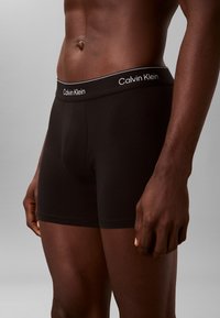 Männlicher Oberkörper trägt schwarze Calvin Klein Boxershorts mit gebrandetem Bund vor einem einfarbigen grauen Hintergrund.