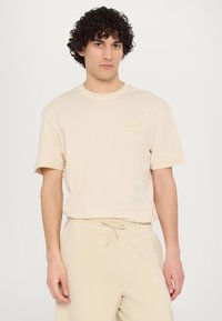 T-shirt en coton beige avec un col rond et des manches courtes, présentant une petite poche à patch sur la poitrine à gauche, accompagnée de pantalons assortis.