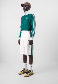 Grön zip-up träningsjacka med vita och gröna ränder, creamfärgade shorts med sidormen, gula sneakers och en brun keps.