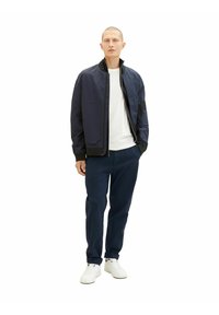 Veste de bombardier marine avec fermeture éclair, associée à un t-shirt blanc et un pantalon marine. Le mannequin porte des baskets blanches. Design simple, sans motifs.