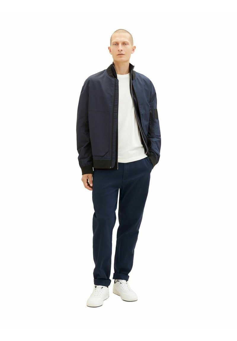 Veste de bombardier marine avec fermeture éclair, associée à un t-shirt blanc et un pantalon marine. Le mannequin porte des baskets blanches. Design simple, sans motifs.