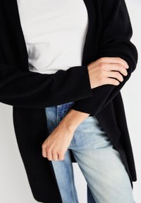Černý cardigan s dlouhými rukávy přes bílý top, který má měkkou texturu a otevřený střih, v kombinaci s světle modrými džíny.