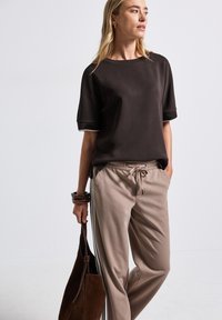 Zwart T-shirt met korte mouwen en witte bies, gecombineerd met beige broek met trekkoord en zijstrepen; model houdt een bruine suède tas vast.