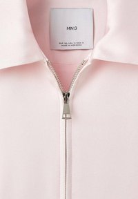 Blouse rose clair avec col, dotée d'une fermeture éclair argentée à l'avant et d'une étiquette d'entretien en tissu à l'intérieur du col.
