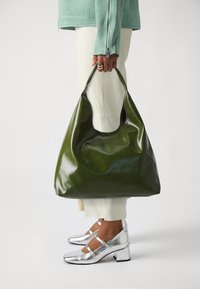 Green glossy oversized handbag with a curved handle. Worn with light-colored pants and metallic silver heels. Smooth texture.