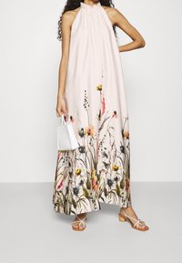 Mujer con un vestido largo sin mangas de color rosa pálido y estampado floral en el dobladillo, sosteniendo un pequeño bolso blanco y usando sandalias blancas con tiras.