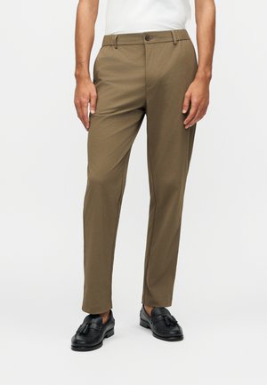 Uomo che indossa pantaloni con taglio a cono verde oliva e mocassini neri con nappine, in piedi davanti a uno sfondo bianco neutro.