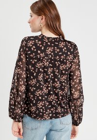 Blouse noire à motif floral avec des manches longues en voile, un col à volants et une coupe ample. Présente des motifs de fleurs roses et crème sur un fond sombre.