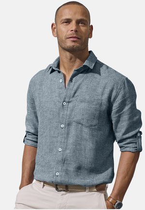 Man with short hair wearing a light blue textured button-up shirt with rolled sleeves, beige pants, and a watch on his left wrist.