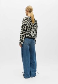Femme avec queue de cheval blonde portant un pull en tricot noir et blanc à motif floral et un jean bleu ample, debout devant un fond blanc uni.