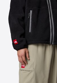 Svart fleecejacka med en asymmetrisk dragkedja och silverstickade detaljer. Bruna cargobyxor med en röd varumärkespatch på fickan.