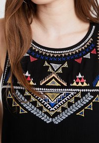 Close-up van een zwarte mouwloze top met kleurrijke geometrische borduursels in rood, blauw, wit, geel en beige rond de halslijn en borst.