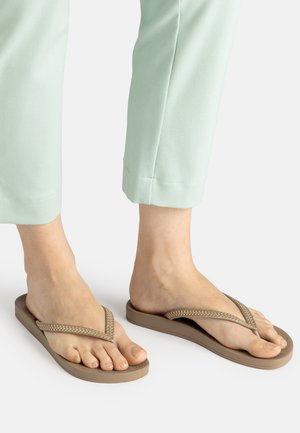 Füße in beigen Flip-Flops mit kleinen dekorativen Nieten, gezeigt mit hellgrünen, gekürzten Hosen vor einem schlichten weißen Hintergrund.