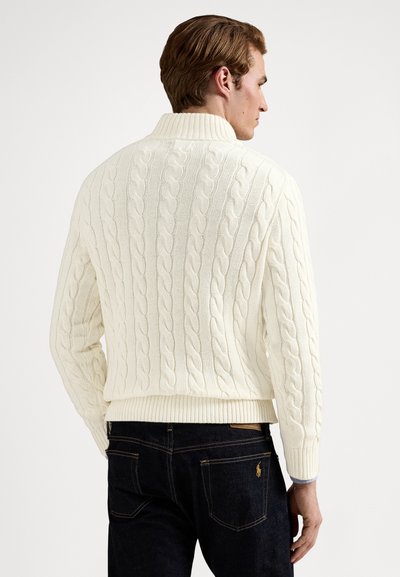 Kabelstrikket genser i kremfarge med ribbet krage og kant, med et teksturert mønster. Kombinert med mørkeblå jeans.