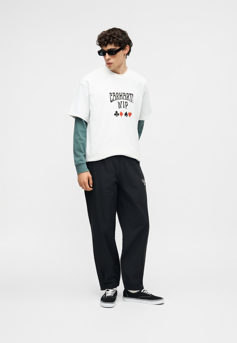 Mann trägt schwarze Sonnenbrille, weißes kurzärmeliges Hemd über grünem Langarmshirt mit Kartenspiel-Symbolen, schwarze Hose und schwarze Sneakers.