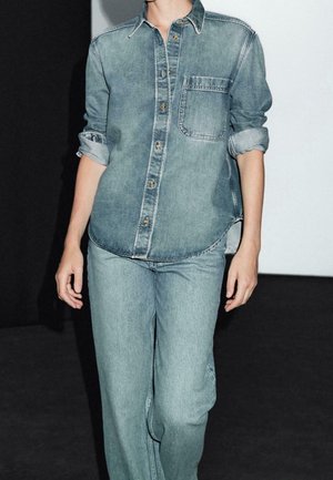 Personne portant une chemise en jean bleu clair avec les manches retroussées et un jean assorti bleu clair sur un fond noir et blanc.