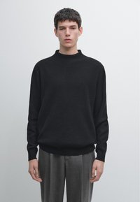 Jeune homme debout devant un fond uni, portant un pull en maille noir et un pantalon gris ajusté, regardant droit devant.