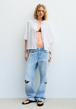 Jeune femme debout contre un mur portant une chemise blanche ouverte, un haut de bikini marron, un jean bleu clair déchiré et des tongs noires.