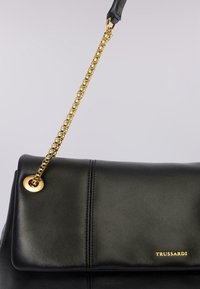 Borsa a mano in pelle nera con patta pieghevole, tracolla in catena dorata, dettagli in hardware dorato e logo impresso. Texture liscia con cuciture visibili.