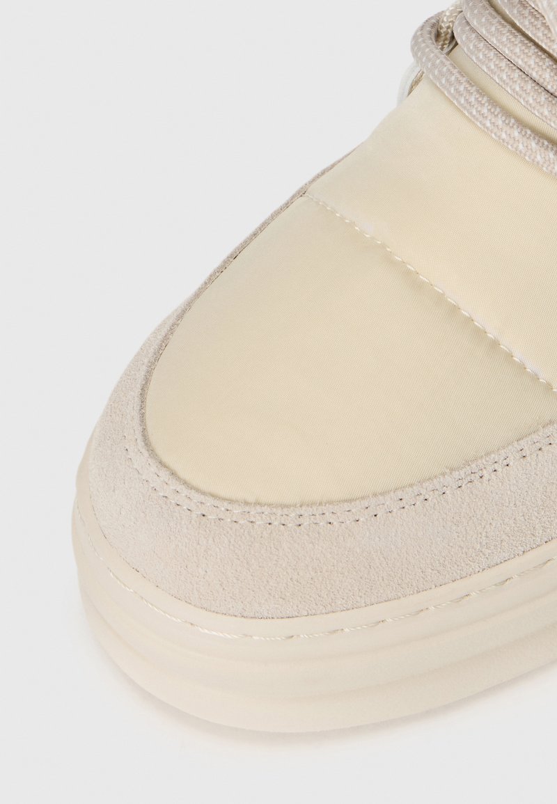 Zapatilla beige con un upper de ante texturizado y nylon suave, forma de puntera redondeada y detalle de costura en contraste.