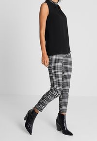 Zwarte mouwloze top met een kralen halslijn, gecombineerd met zwart-witte geruite skinny pants en glanzende zwarte enkellaarsjes met een blokhak.
