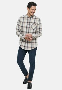 Chemise à carreaux beige, bleu et marron, dotée d'une poche poitrine et d'un ourlet incurvé ; associée à un jean foncé et des mocassins noirs.