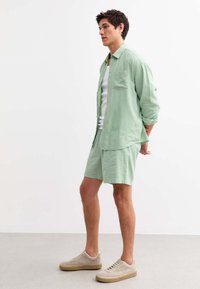 Set di camicia e pantaloni corti in lino verde menta, caratterizzato da una vestibilità ampia, una tasca singola sul petto e abbinato a sneakers beige. Texture morbida.
