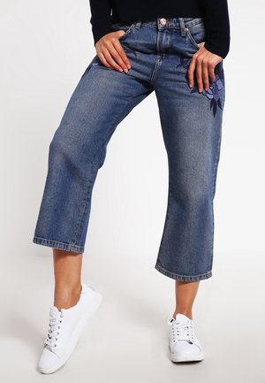Frau trägt kurze blaue Jeans mit lila Blumenstickerei, kombiniert mit weißen Sneakers, steht mit Händen in der Hüfte.
