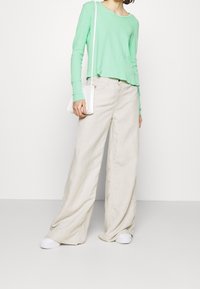 Tyrkysově zelený top s dlouhým rukávem v kombinaci s volnými béžovými lněnými kalhotami. Bělé tenisky a malá bílá crossbody kabelka dolaďují celkový vzhled.